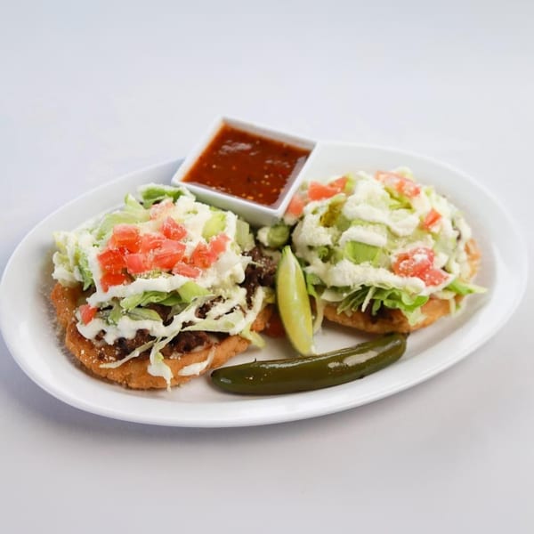Two Mexican sopes topped with meat, lettuce, cheese, and tomatoes, with salsa and a jalapeño.