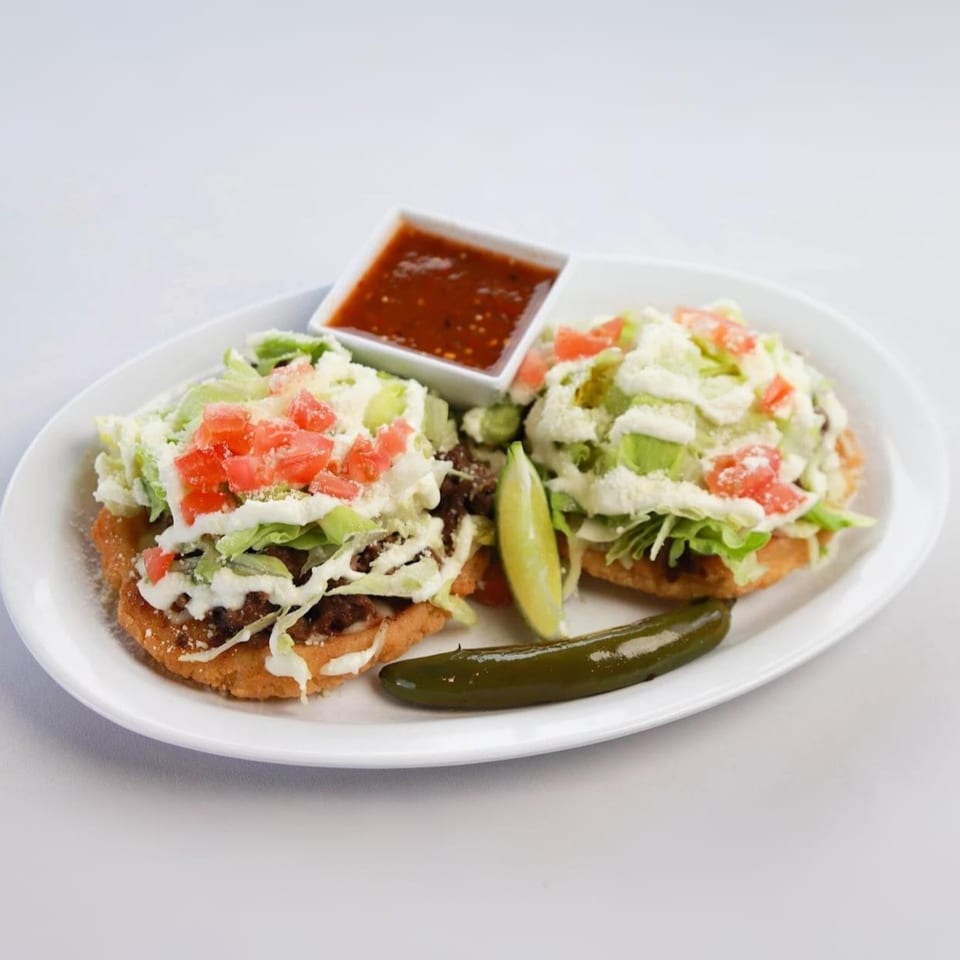 Two Mexican sopes topped with meat, lettuce, cheese, and tomatoes, with salsa and a jalapeño.
