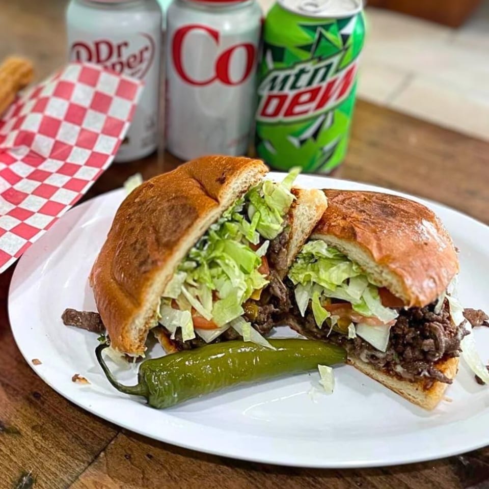A Mexican torta sandwich, cut in half, with a green chili and three soda cans.