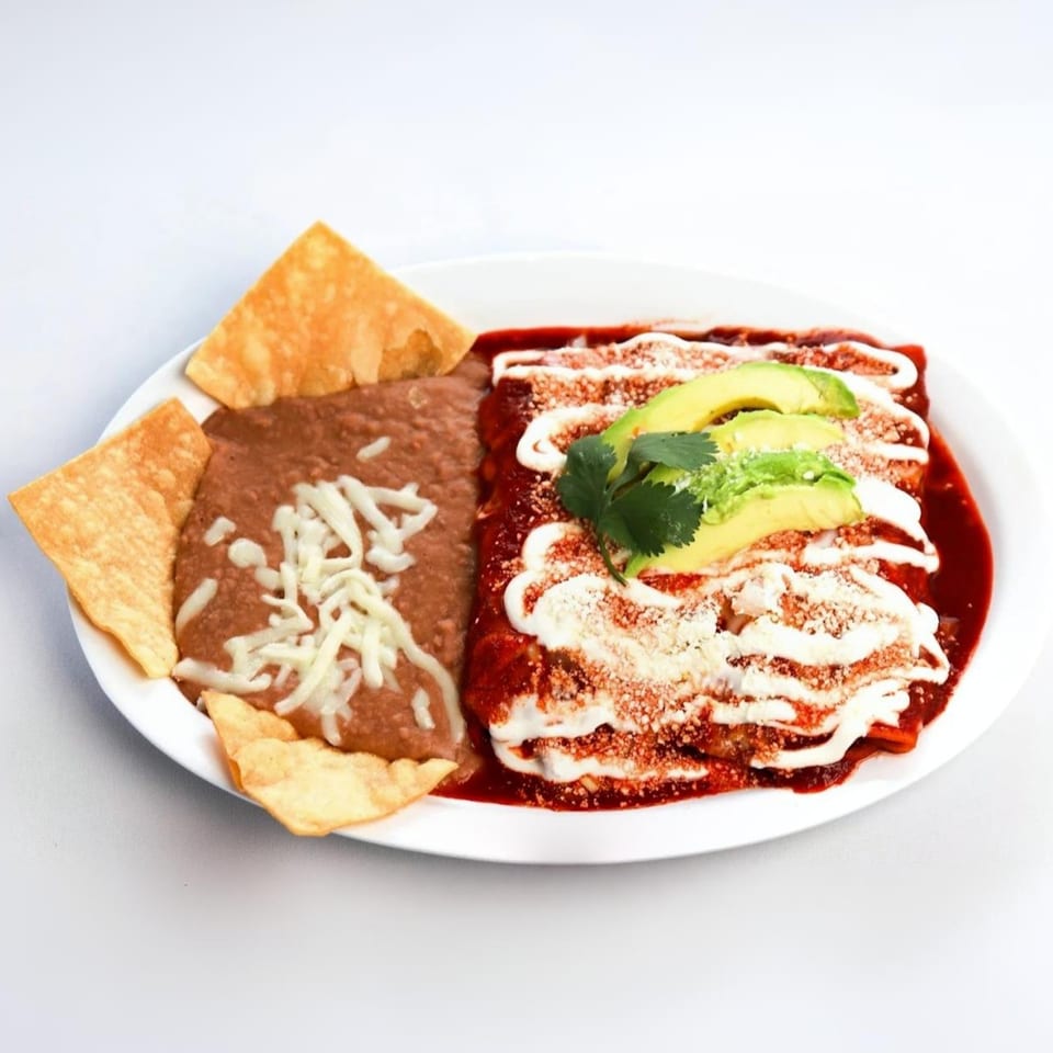 A plate of Mexican enchiladas with red sauce, sour cream, avocado, cilantro, refried beans, and chips.