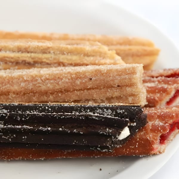 A close-up of various churros, including classic, chocolate, and fruit-filled, on a white plate.
