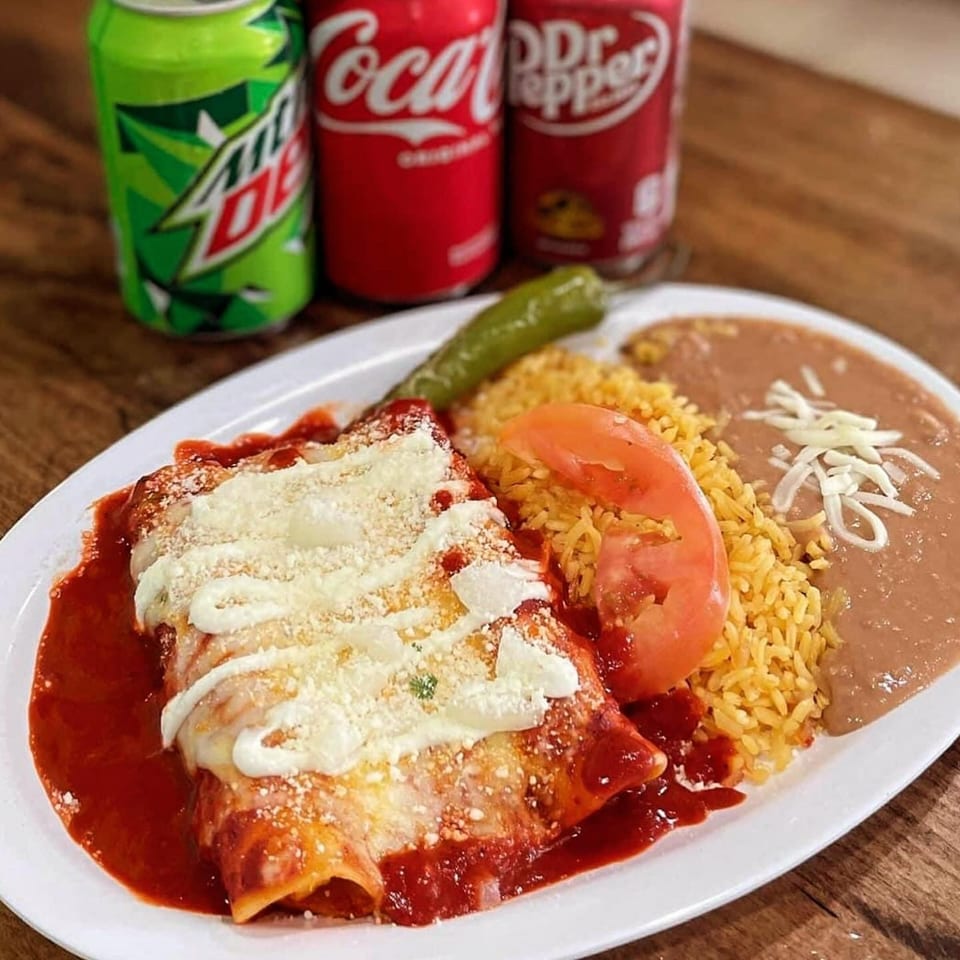 Two enchiladas with red sauce, rice, beans, and a green chili, with three soda cans in the background.