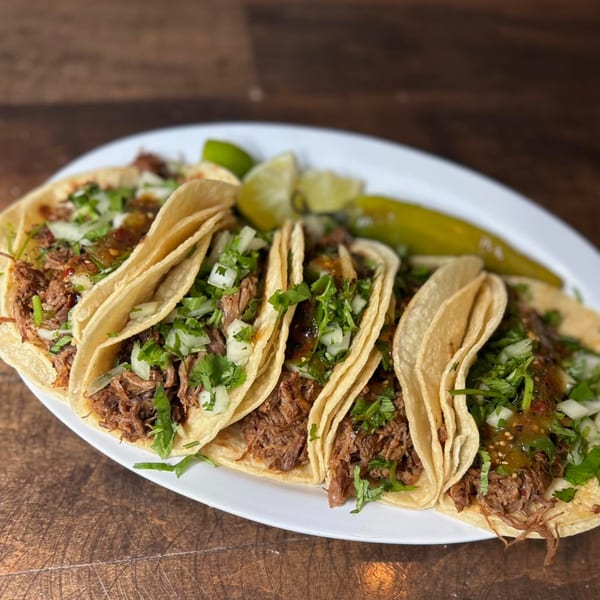 Five Mexican street tacos with shredded meat, cilantro, onions, and salsa, served with lime and a pepper.